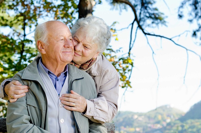 An elderly couple, one person hugging the other from behind, they are smiling.