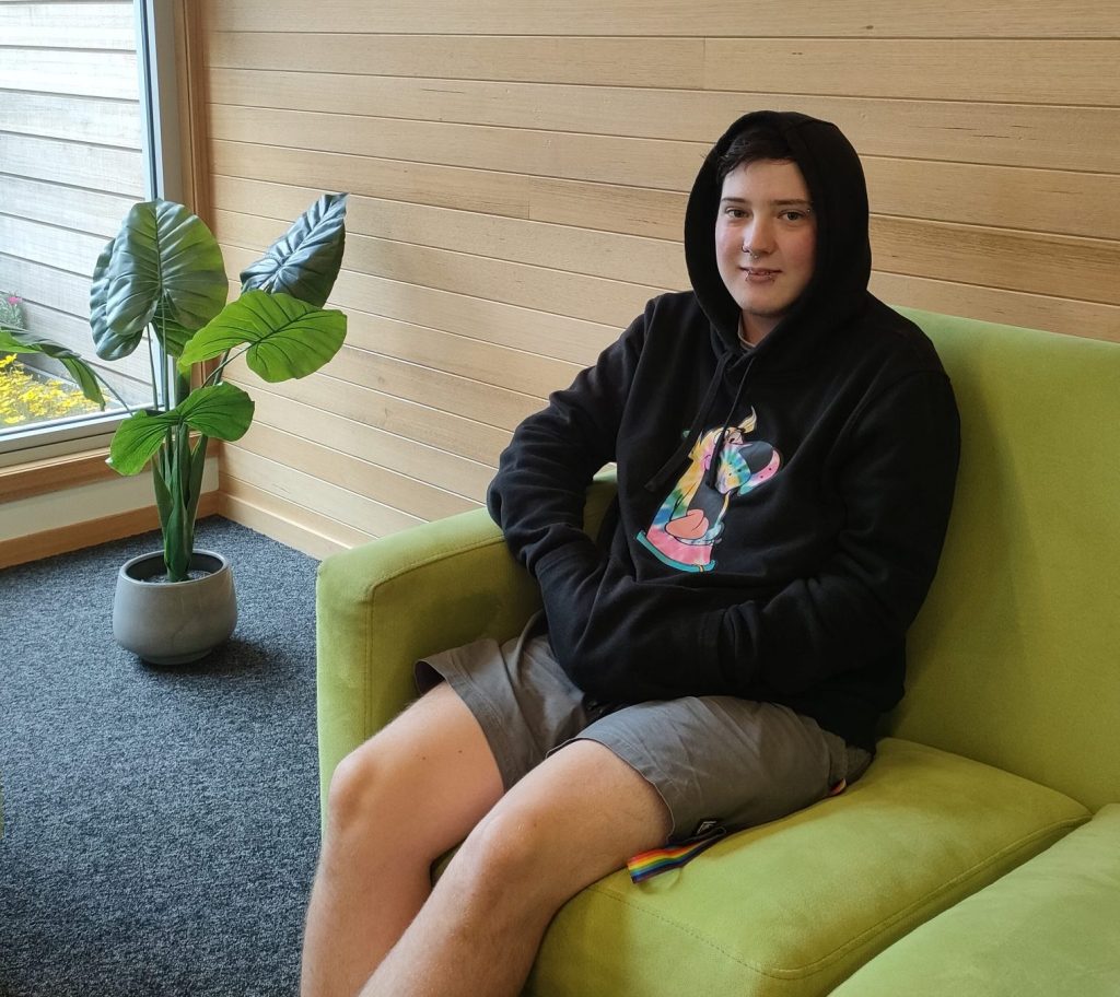 Parker, Eveline House resident is sitting on a green couch wearing a hoodie and a pair of shorts. He is smiling.