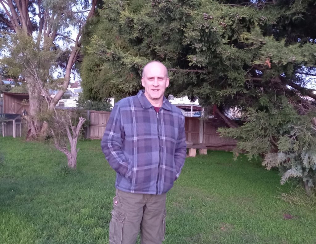 person standing in a garden with pants and a jumper on, he is smiling with his hands in his pockets.