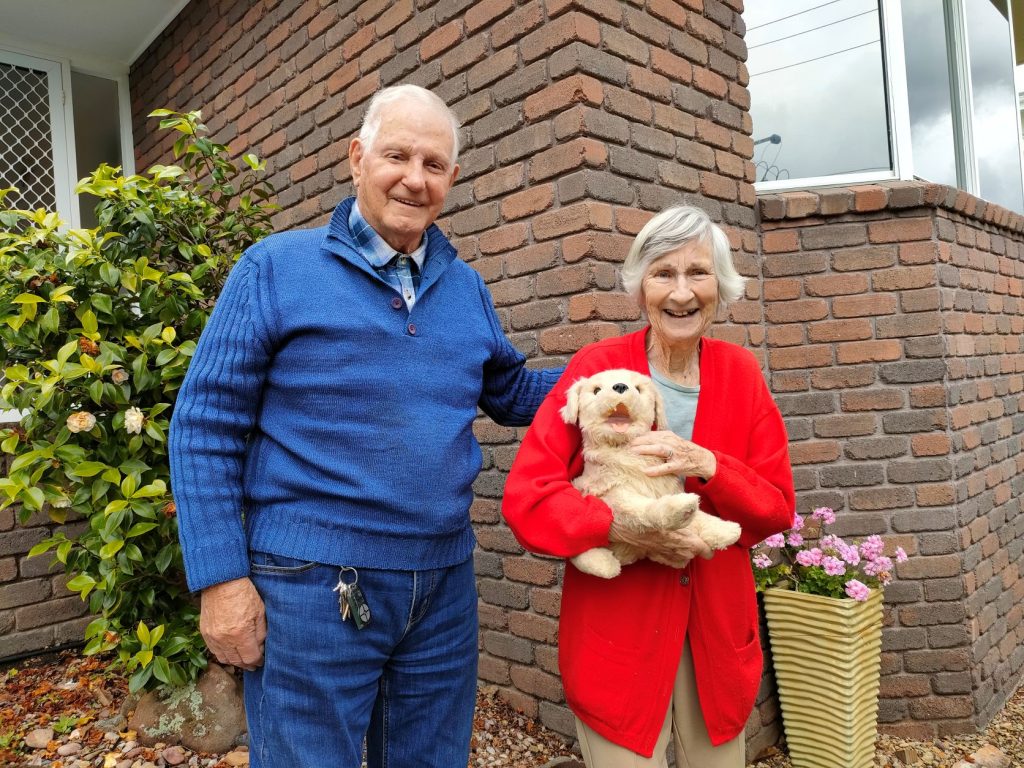 Clients Brian and Judy standing together oustide their house. Judy is holding a soft toy that looks like a dog. They are both smiling.