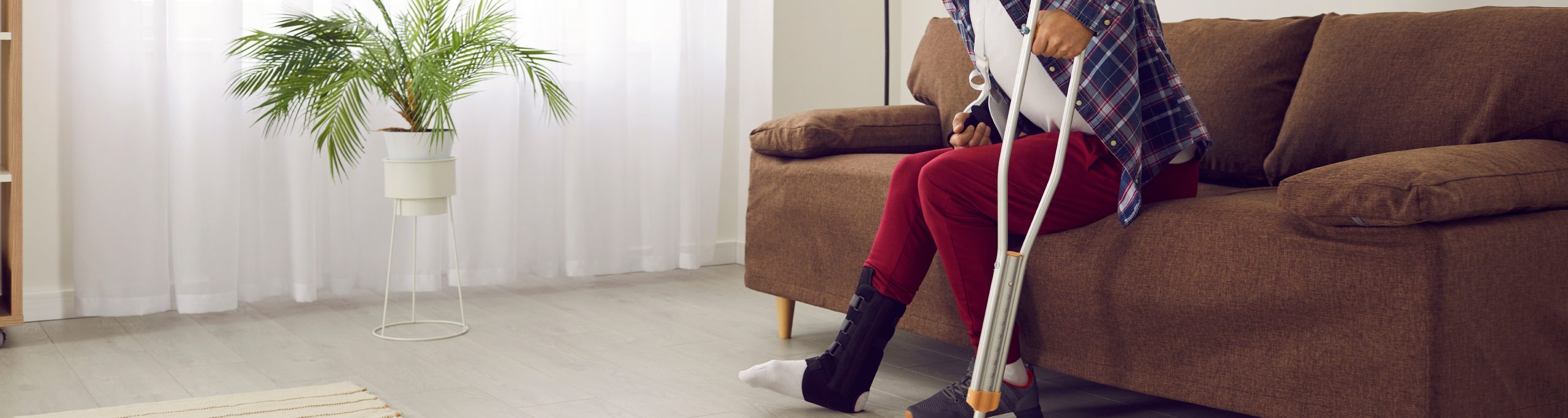 person sitting on a couch with a plaster cast on their leg and holding a crutch. Indicates they have had an accident.