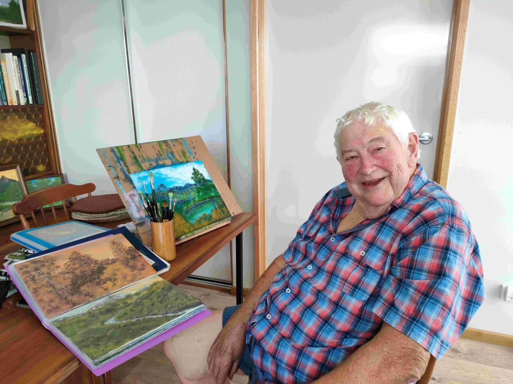 Lloyd OMalley sitting at his desk smiling. His landscape paintings are sitting beside him.