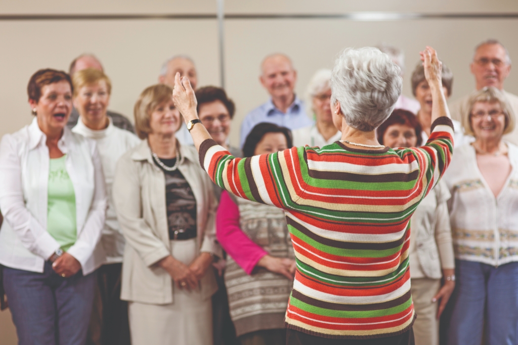 A lady standing up in front of a group of people which appear to be a choir, singing. She has her hands up in the air. The people singing look happy and to be enjoying it. 